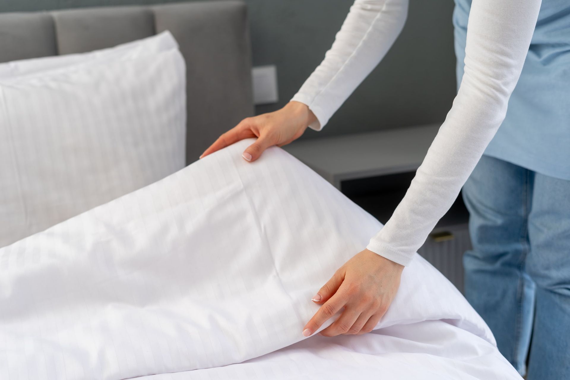 Hands smoothing a white duvet on a bed with a white pillow and grey headboard.