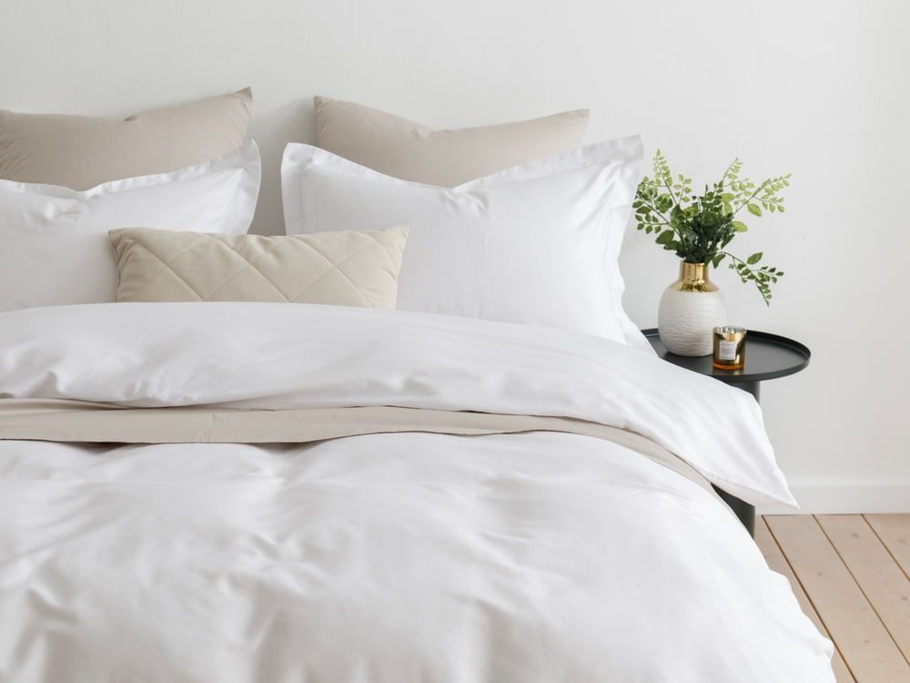 White bedding on a bed with neutral pillows. A side table holds a vase and small objects.