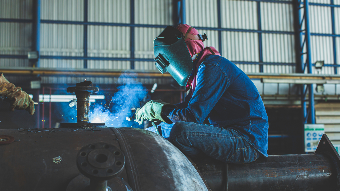 Welder in blue work clothes and helmet, using a welding torch on a large metal pipe in a factory setting.
