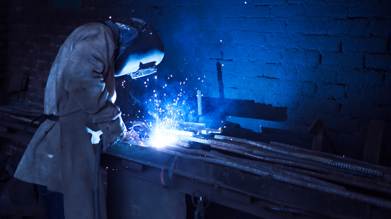 Welder wearing protective gear, welding metal in a dark workshop, sparks flying amidst the blue light.