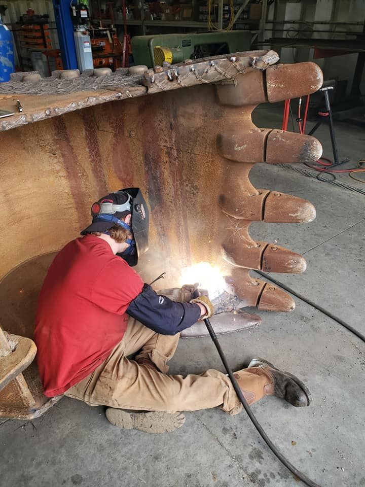 A man is welding a large excavator bucket in a garage.