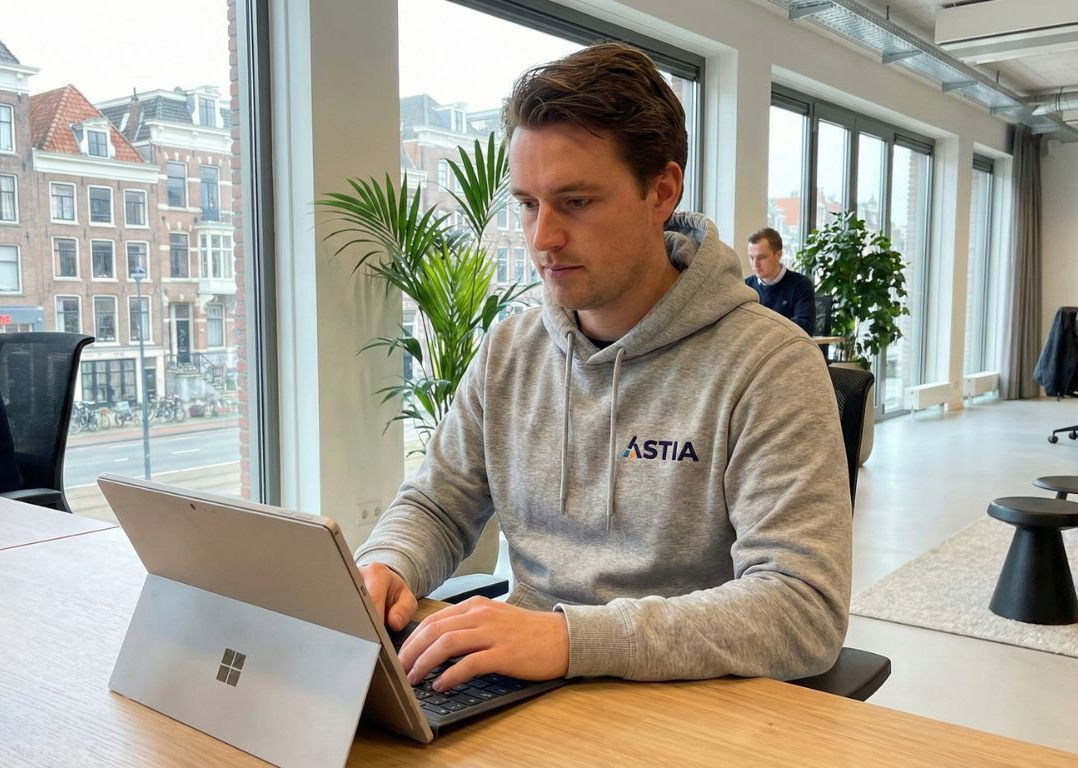 Een man werkt op een laptop in een licht kantoor; op de achtergrond zijn planten, ramen en andere mensen te zien.