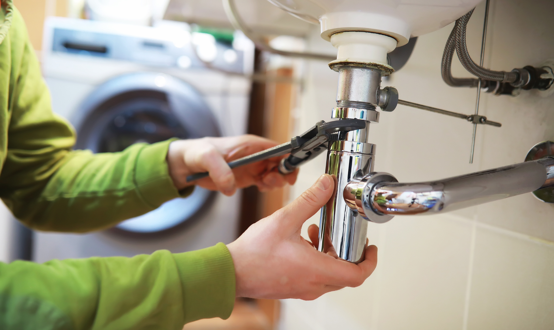 Person using a wrench to work on plumbing under a sink; a washing machine is in the background.