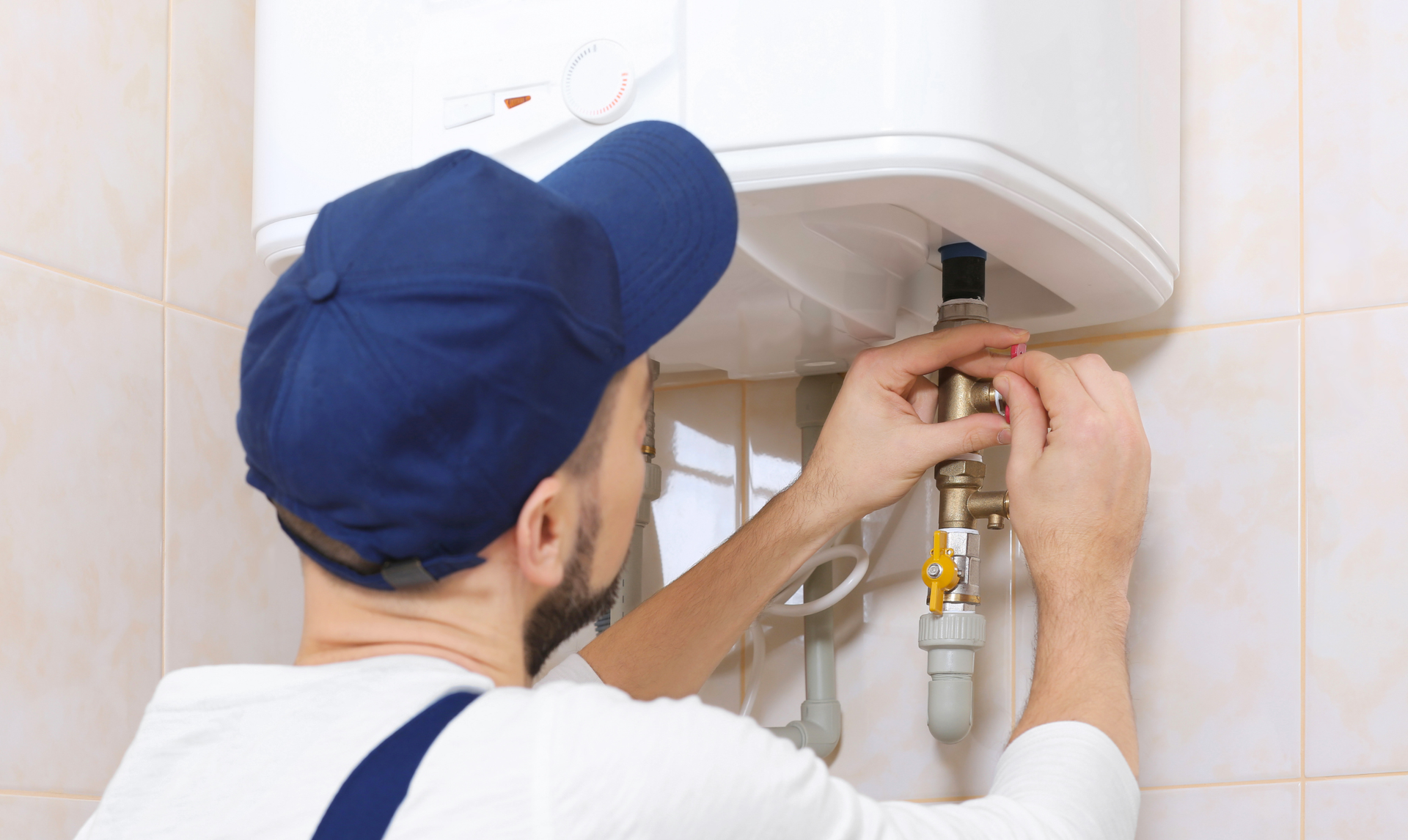 Plumber in blue cap and coveralls adjusting plumbing on a white hot water tank in a tiled bathroom.