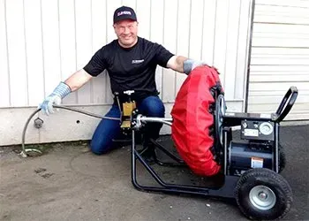 Man kneels beside a drain cleaning machine, holding the cable near a building's pipe.