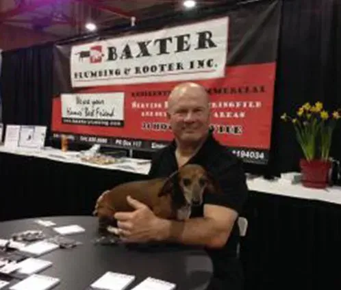 Man holding dachshund at Baxter Plumbing & Rooter Inc. booth. Black and red banner.