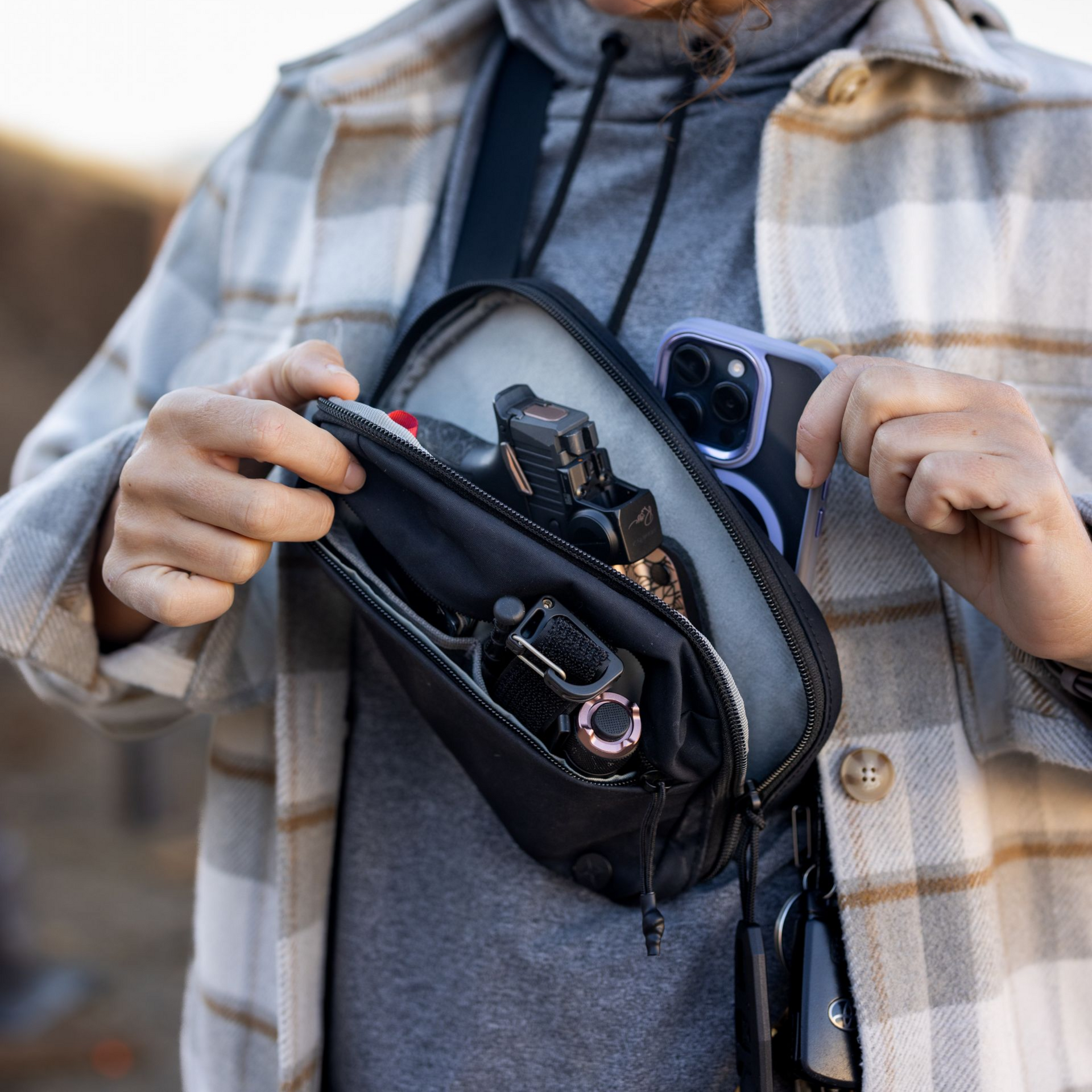 A person wears a black sling bag across their chest, showing organized compartments containing a phone and tools.
