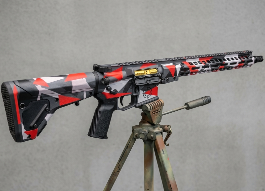 A rifle with a red, black, and white camo finish mounted on a tripod against a grey background.