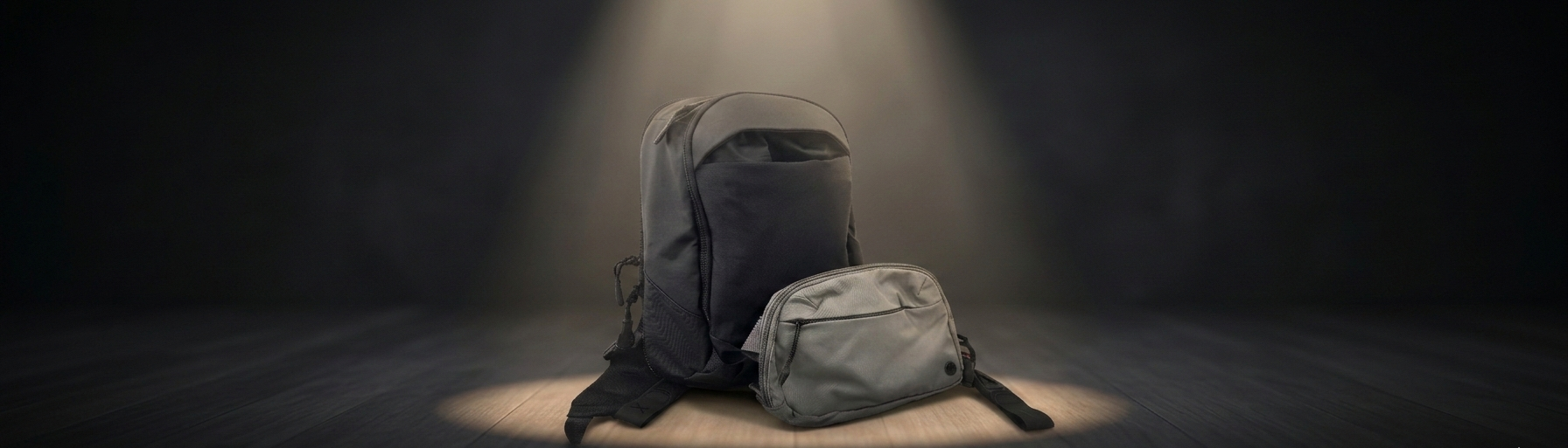 A gray backpack and matching sling bag sitting together on a dark, spotlighted surface.