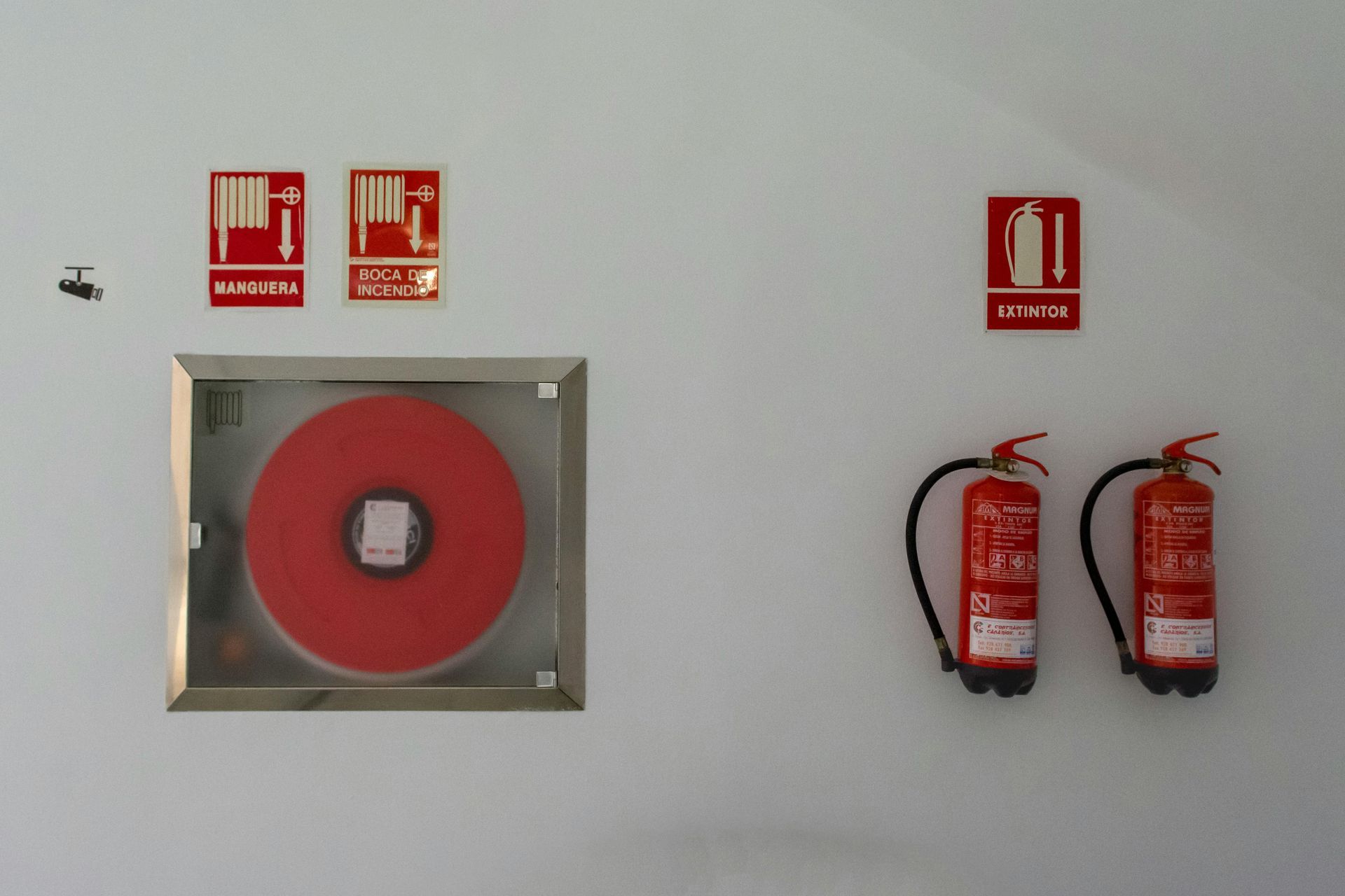 A fire hose reel in a glass cabinet and two red fire extinguishers mounted on a white wall with corresponding signs above.