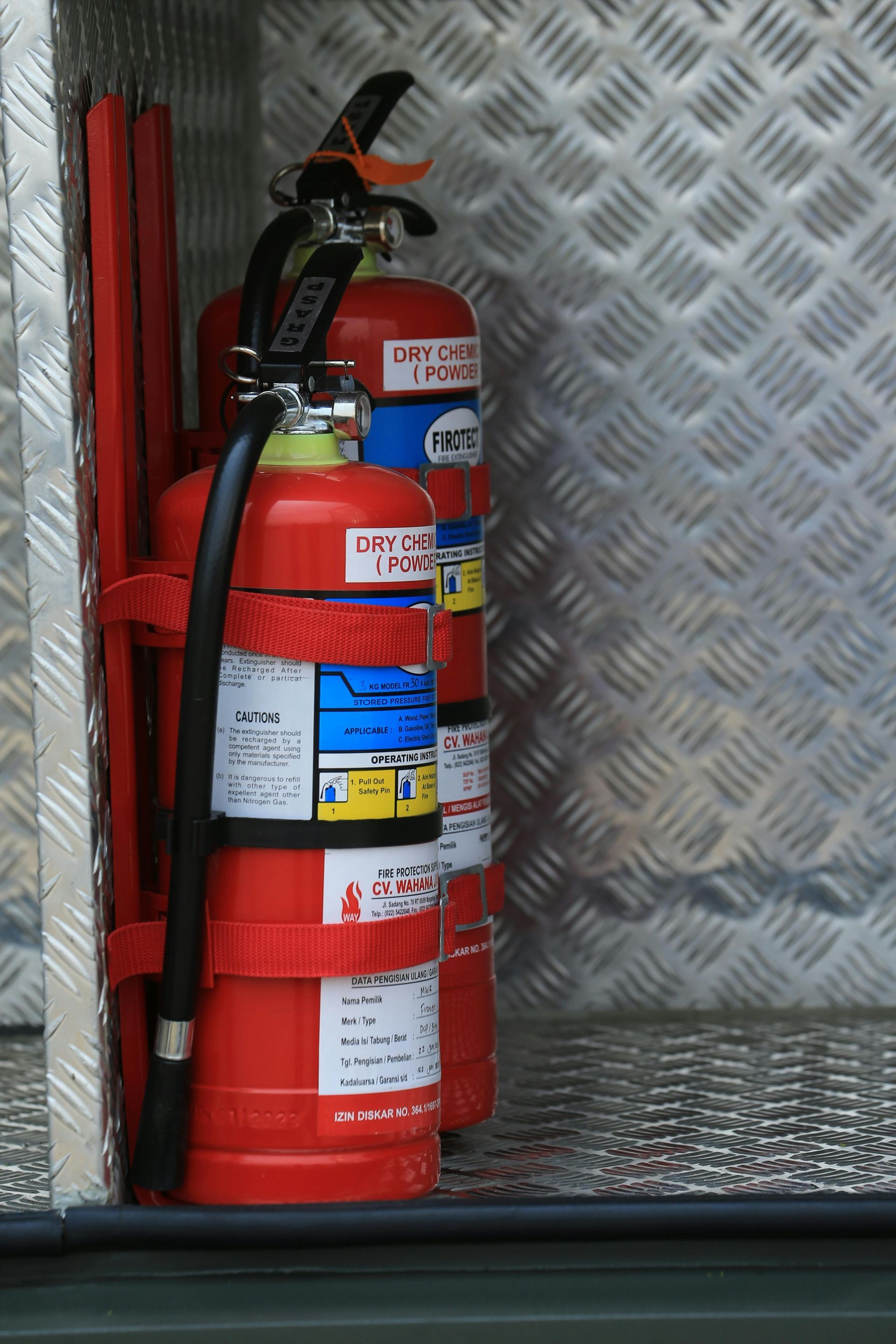 Two red fire extinguishers secured with straps inside a metal compartment with a diamond-plate pattern.
