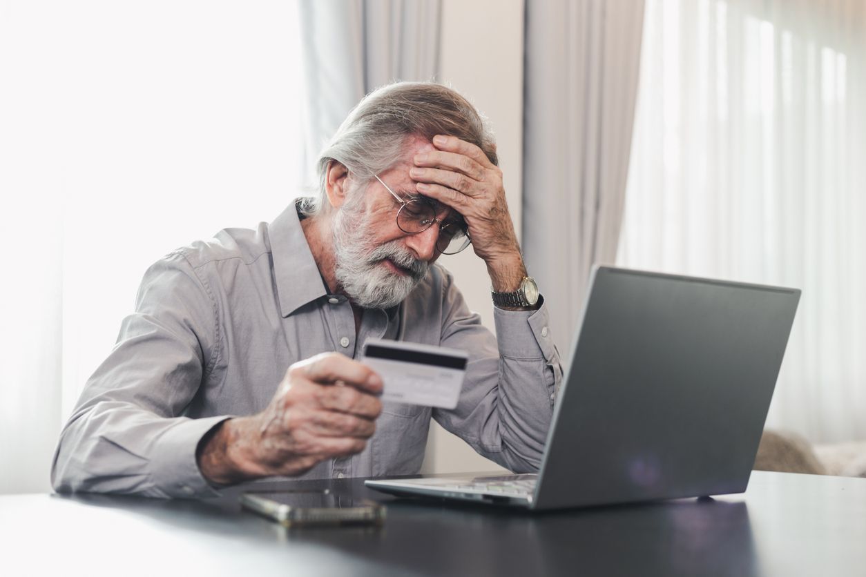 Man looking stressed, holding credit card, using a laptop at a table indoors.