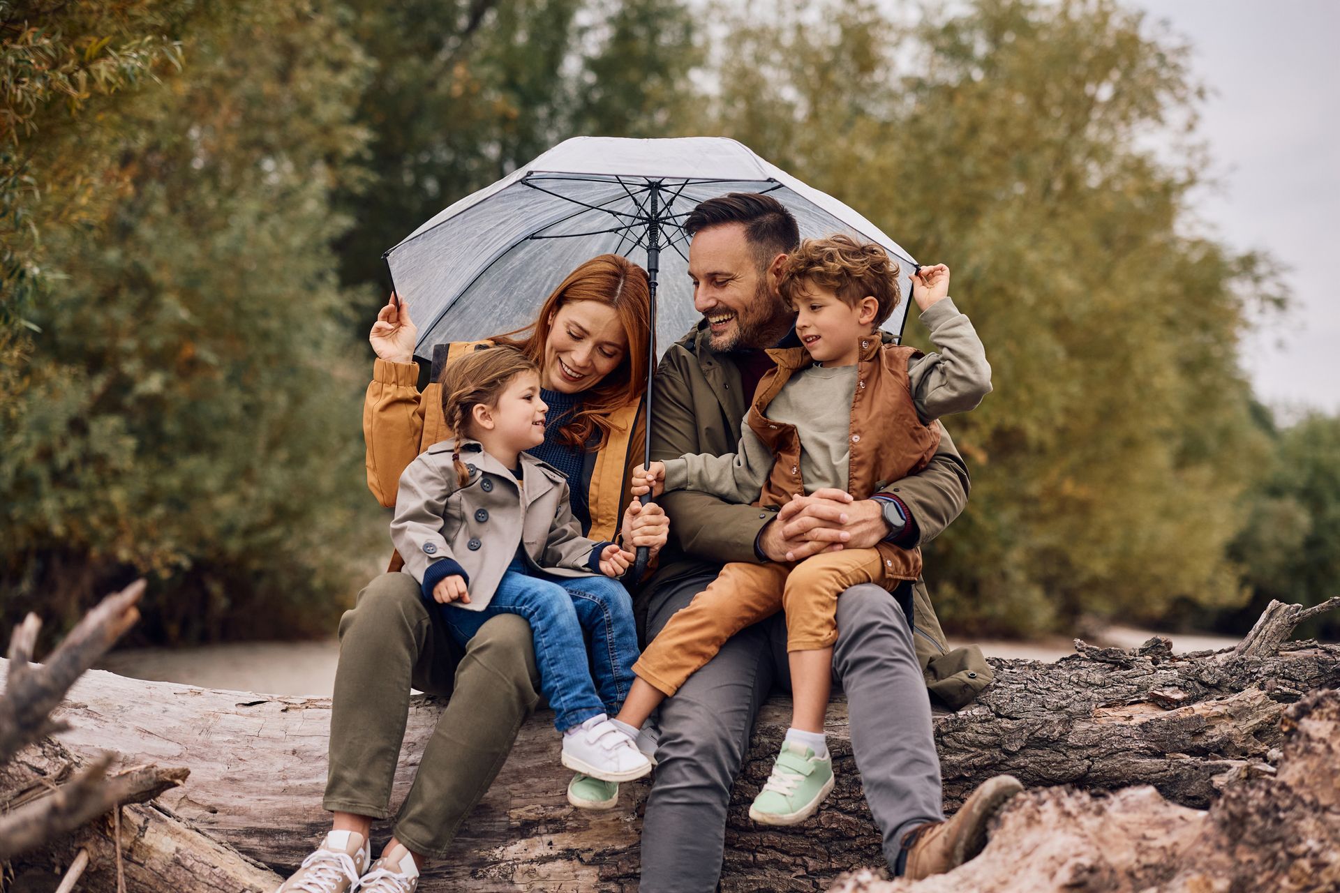 Family of four under umbrella sitting on a log outdoors, smiling.