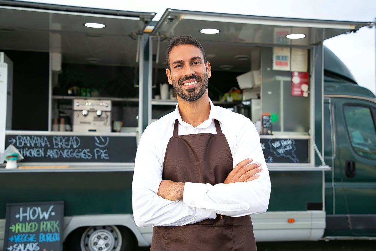 A smiling person wearing a white shirt and brown apron stands with arms crossed in front of a food truck.