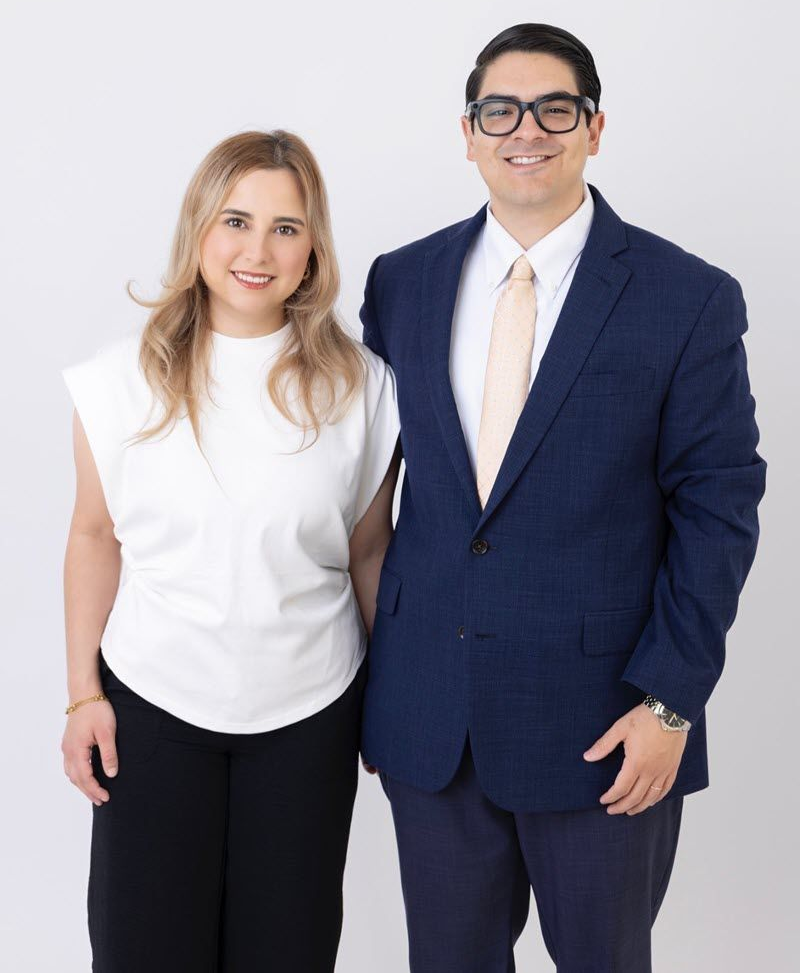 A woman and a man pose together; the man wears a suit and glasses, and the woman wears a white top and black pants.