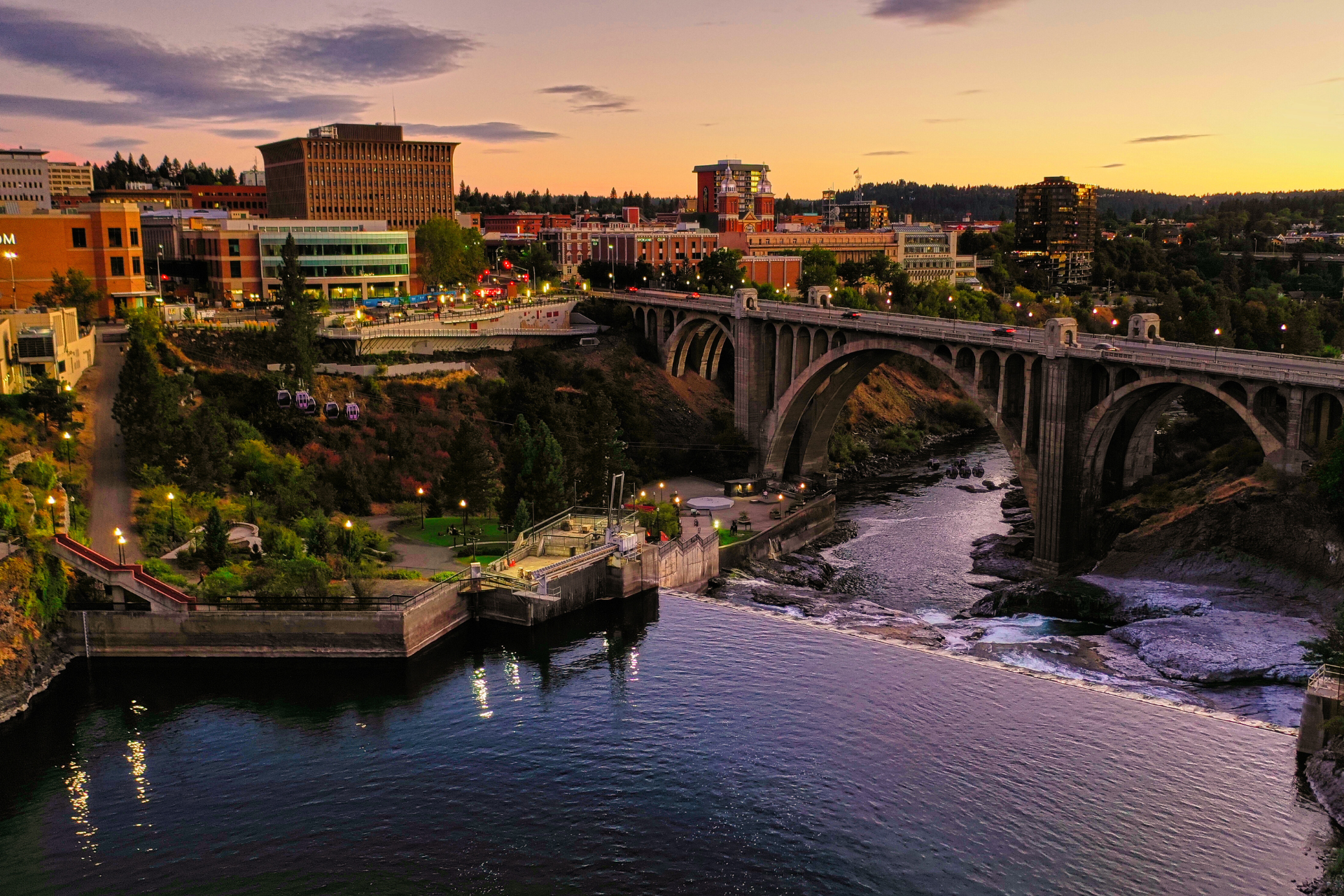 Sunset over Spokane, Washington, with a river flowing under a large concrete arch bridge. City buildings line the banks.