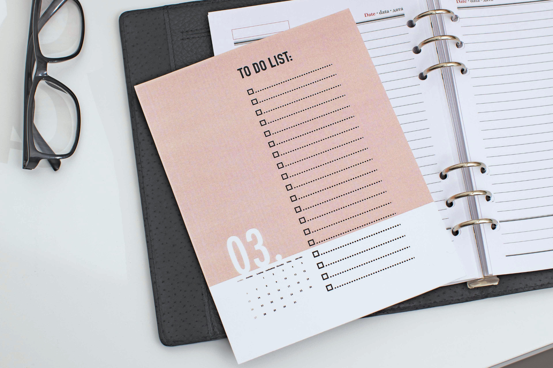 To-do list on pink paper in a binder, with glasses on a white desk.
