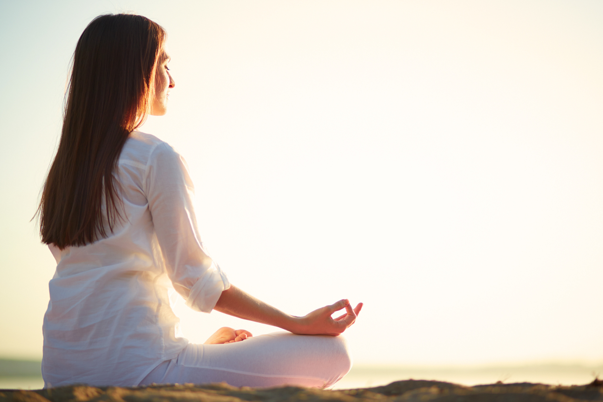 Woman in white clothes meditating with eyes closed, outdoors in bright sunlight.