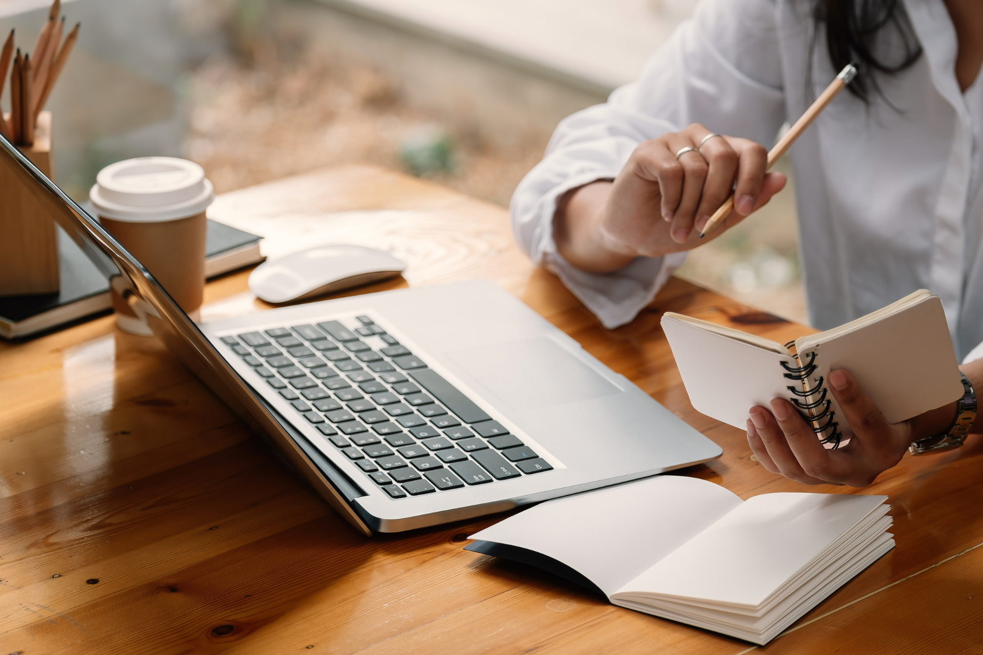 Woman working on a laptop, taking notes in a notebook at a wooden desk. A coffee cup and mouse are also on the desk.