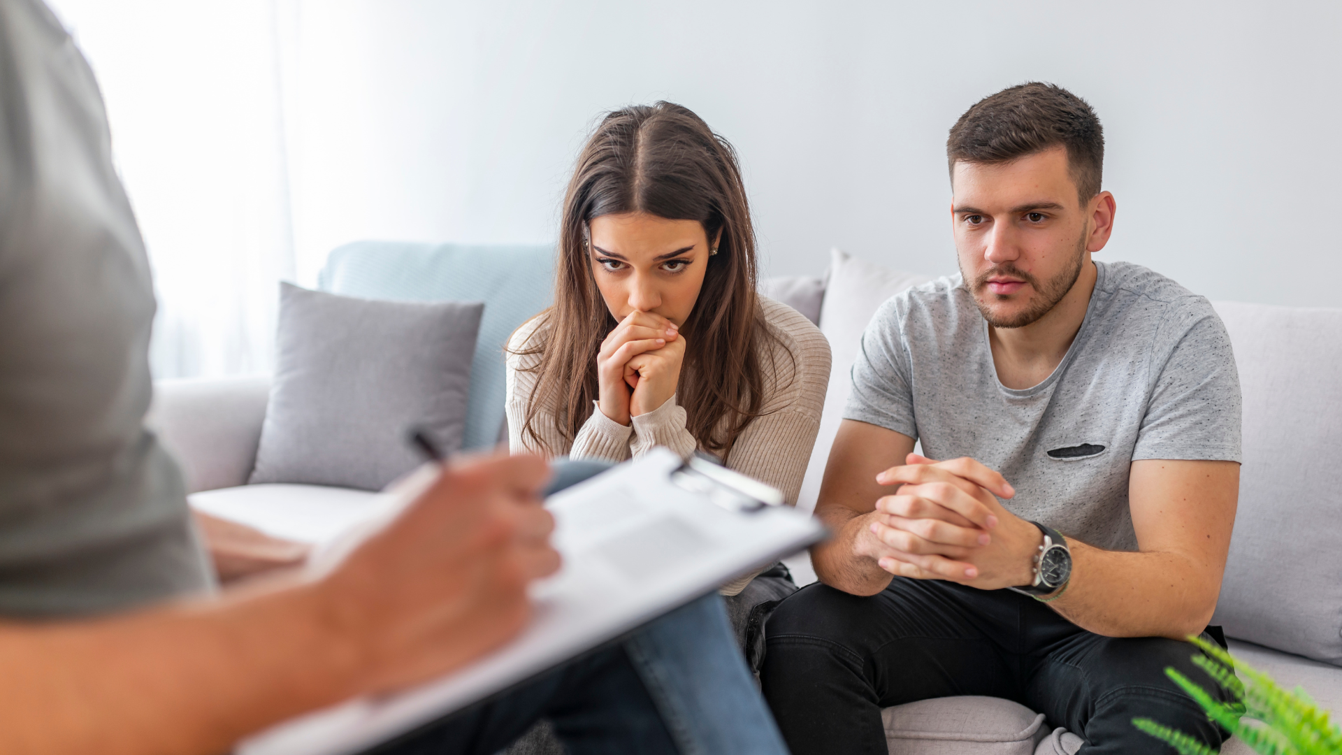 Couple on a couch with a therapist taking notes; all appear worried, light room.