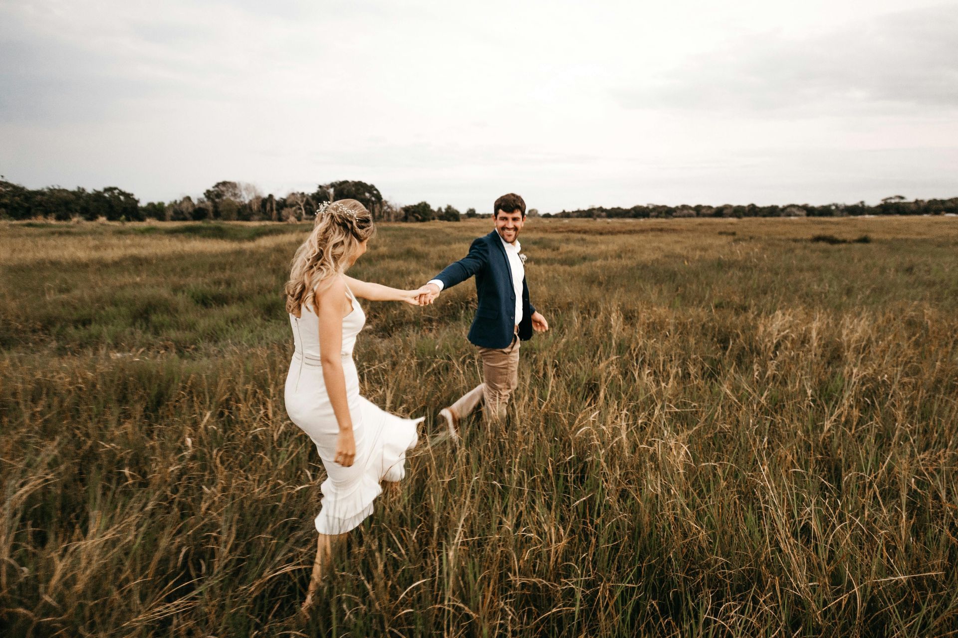 A bride and groom are running through a field holding hands.