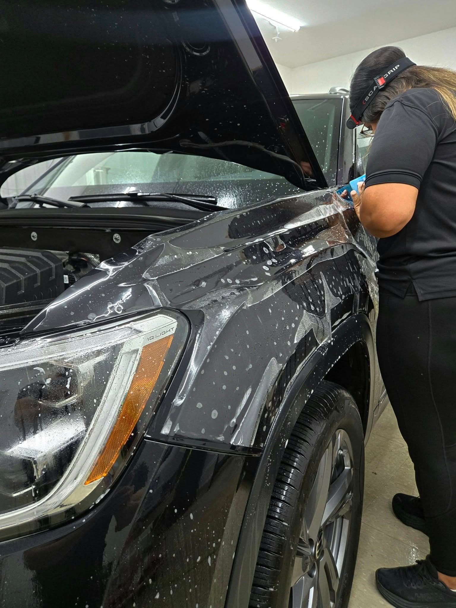 Person applying protective film to a black car's hood in a workshop.