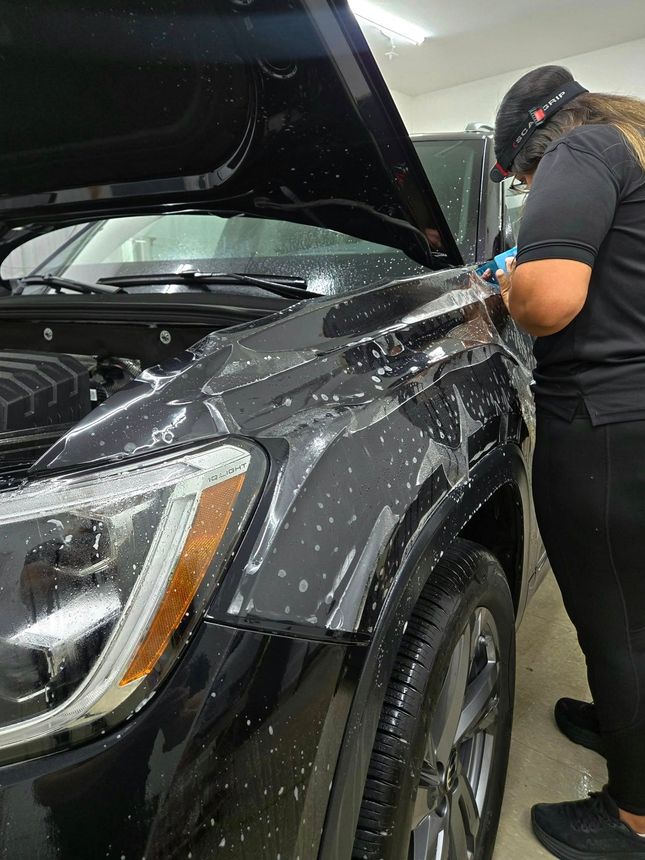 Person applying protective film to a black car's hood in a workshop.