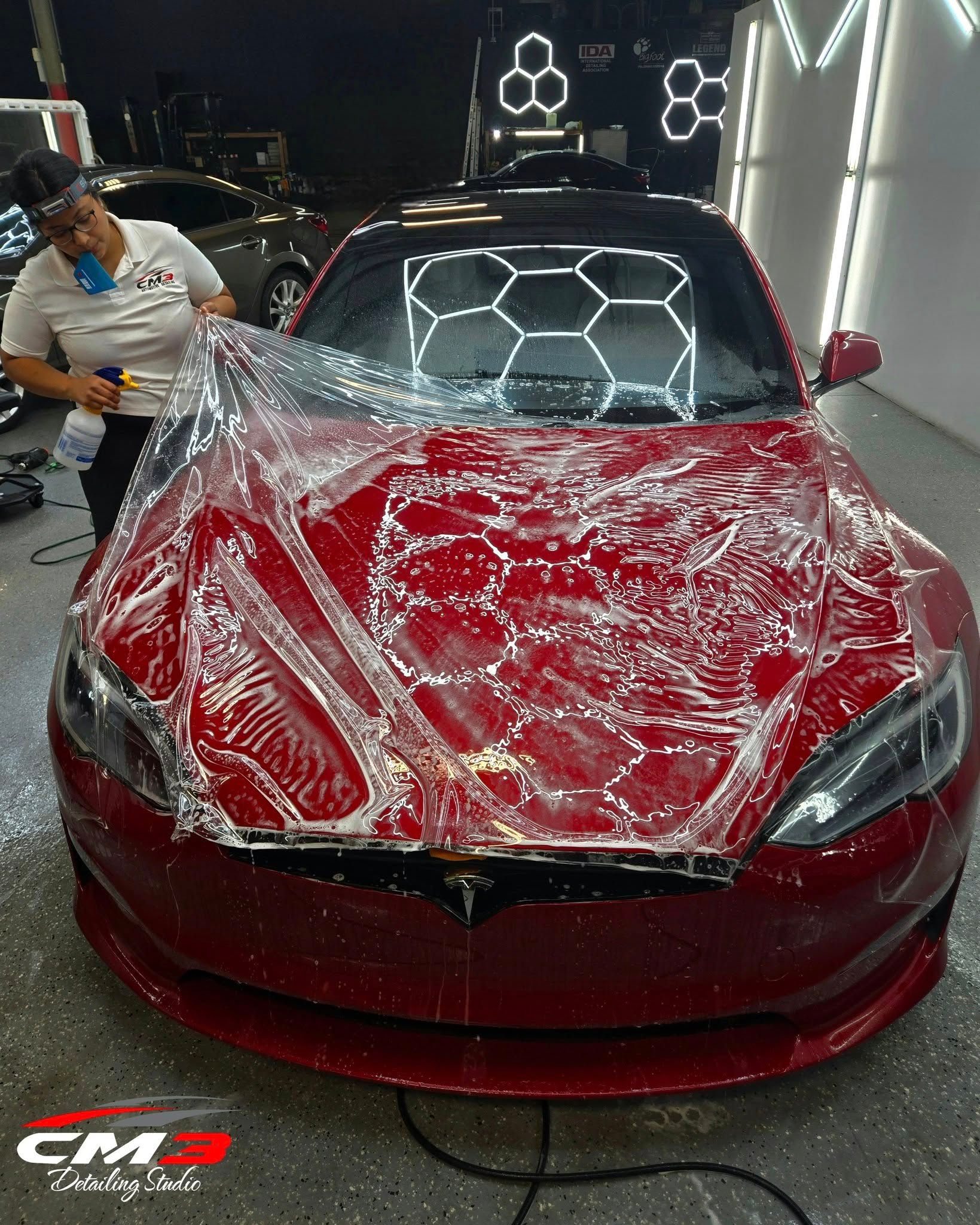 Person applying protective film to a red Tesla. Shop interior with bright lights.