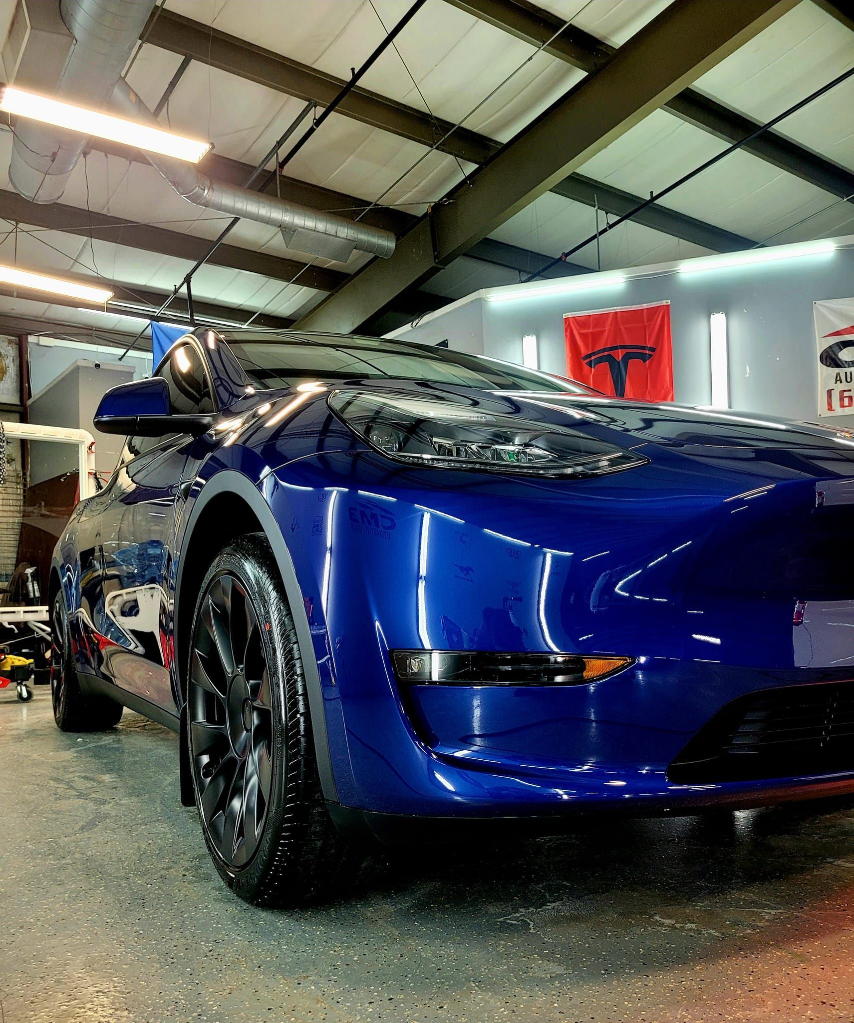 Blue Tesla car parked inside a well-lit shop, glossy finish, with a Tesla flag in the background.