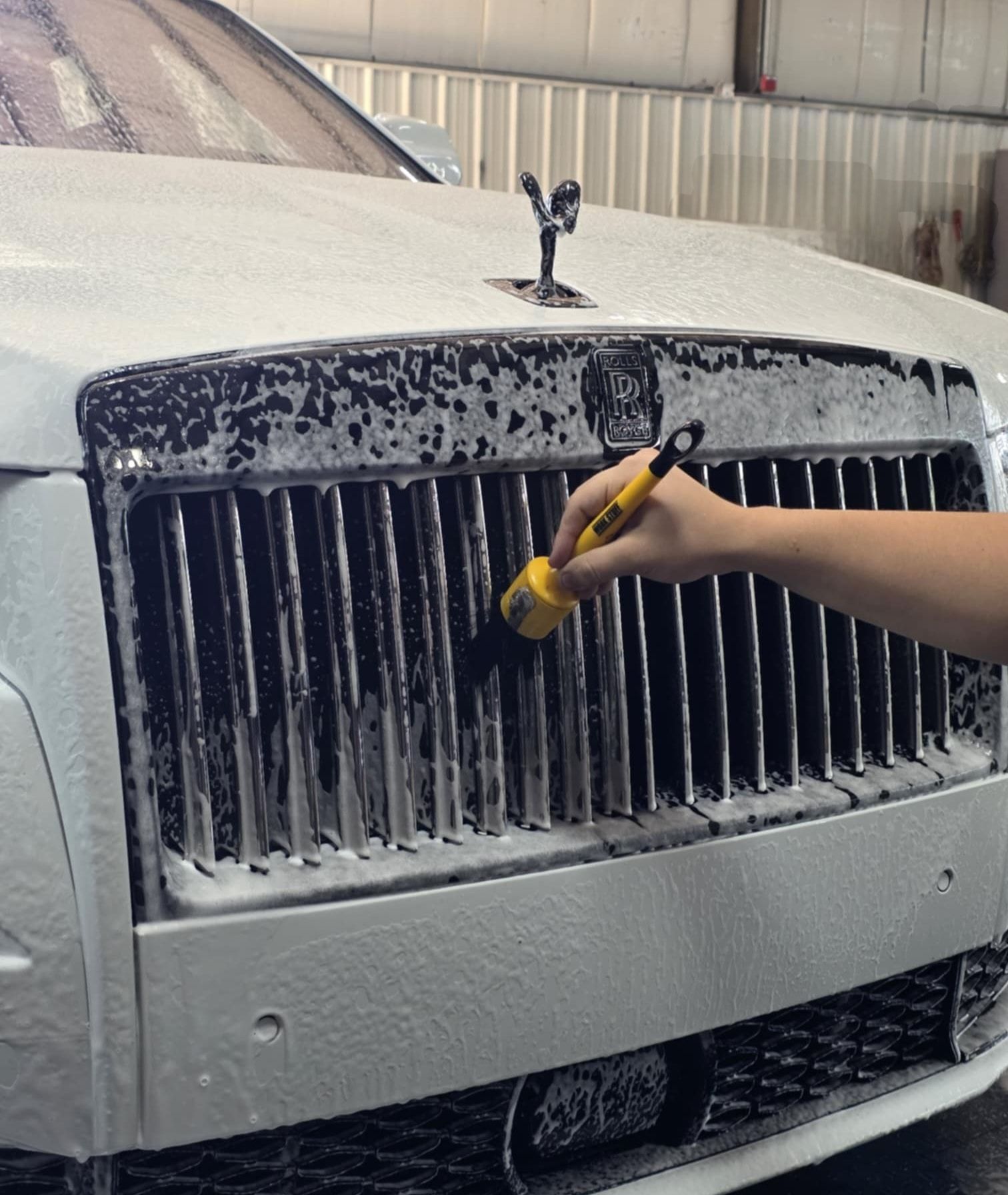 A person cleans a white Rolls-Royce grill with a brush covered in soapy foam.