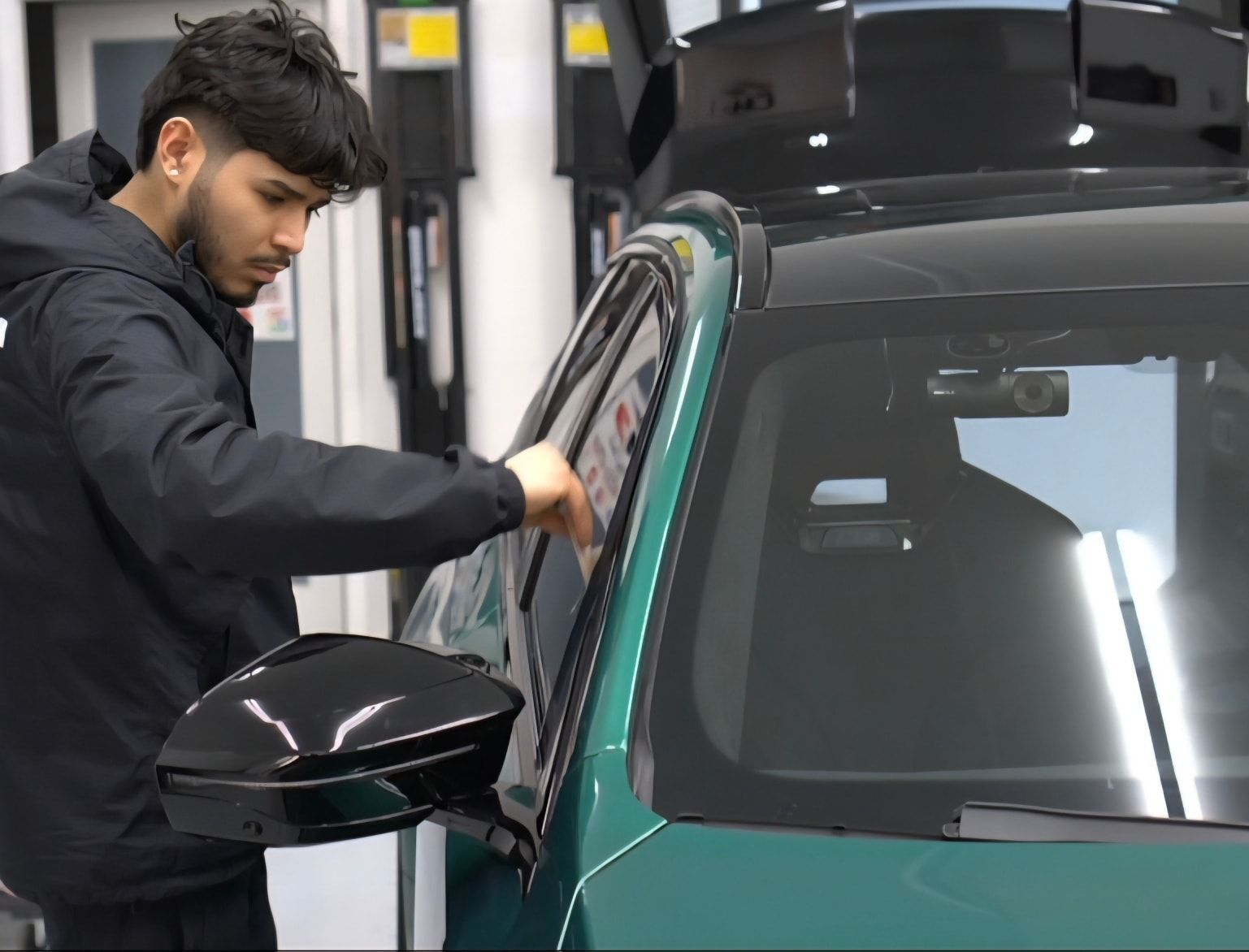 Man in black jacket leaning toward a green car, touching the windshield frame.