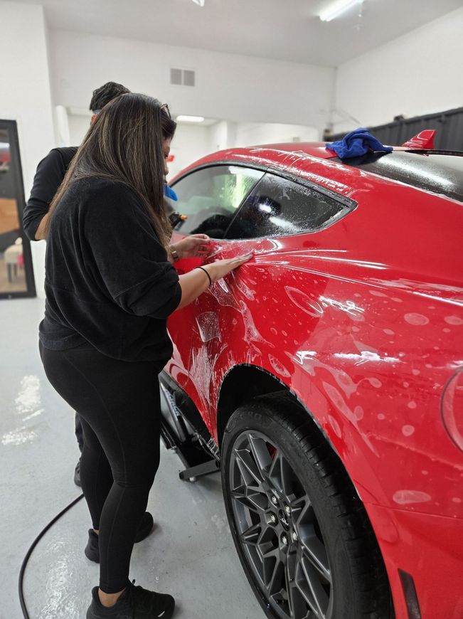 Two people applying film to a red car in a garage.