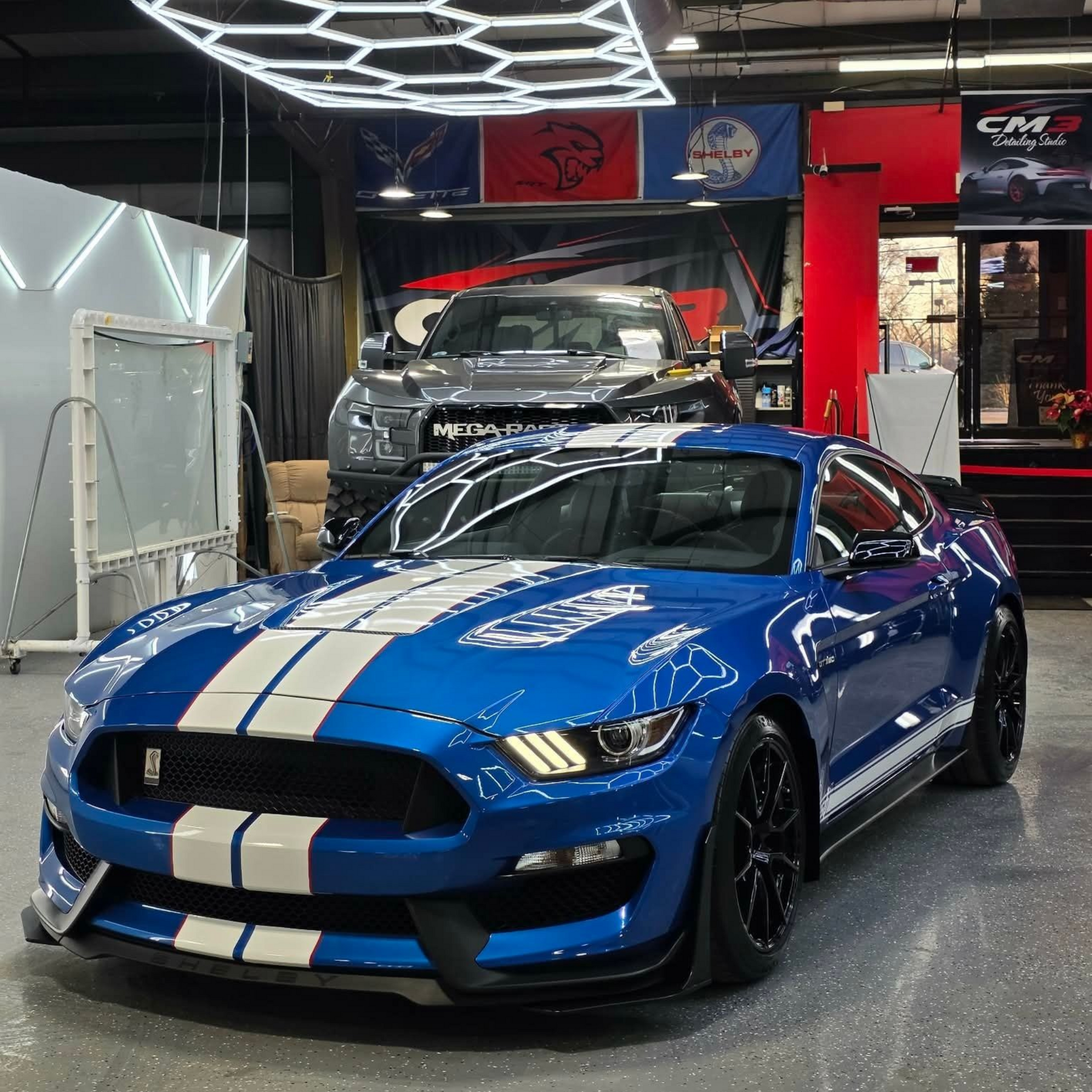 Blue Ford Mustang Shelby GT350 with white stripes, parked in a car detailing shop, with a pickup truck in the background.