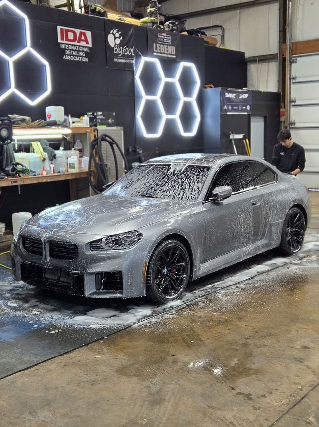 A grey BMW coupe covered in soap suds, being washed in a shop; person in the background.