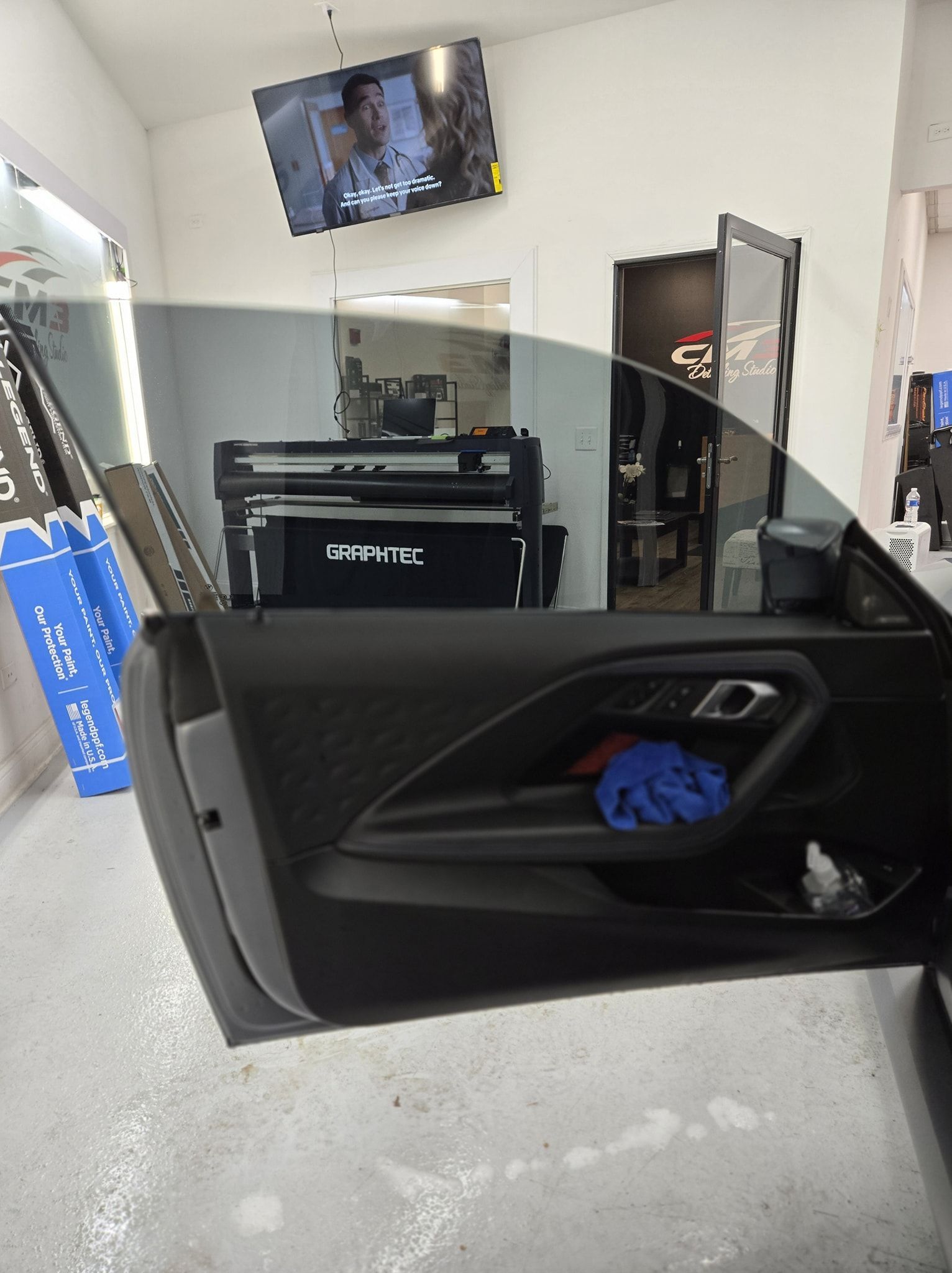 Car door open in a shop, showing interior and tinted window. Tinting equipment visible in the background.