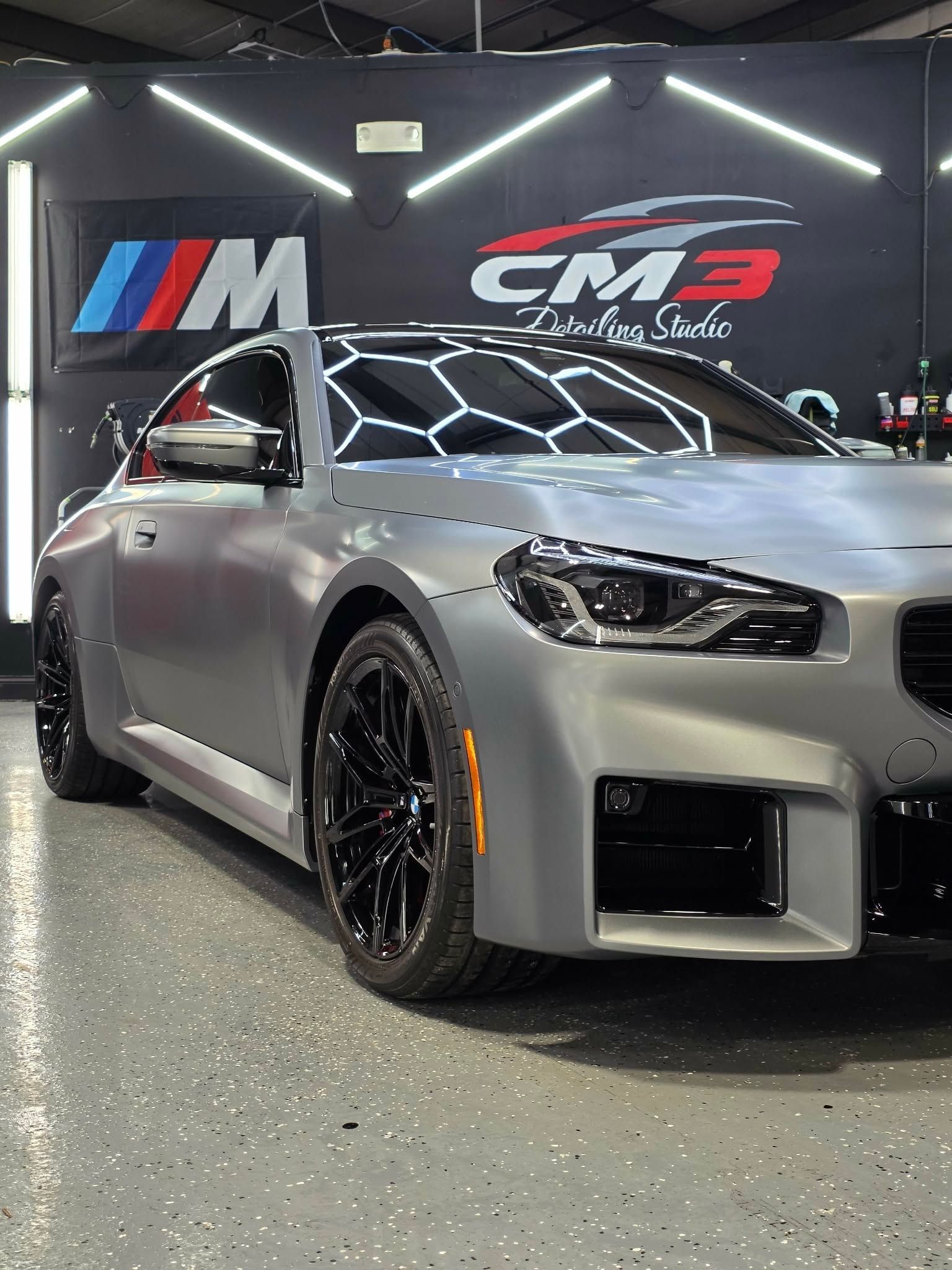 A gray BMW coupe in a car detailing studio, black wheels, under bright lights, CM3 logo.