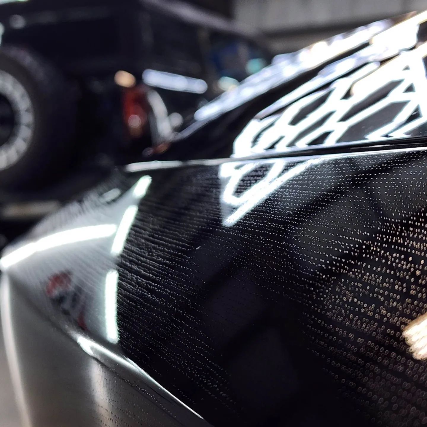 Close-up of a glossy, textured black car surface with reflective highlights, likely carbon fiber.
