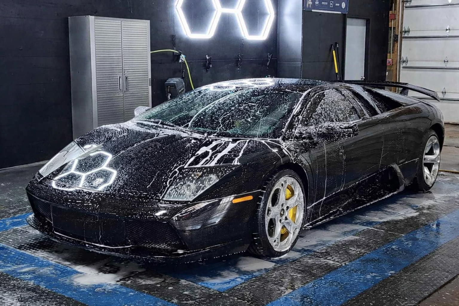 A sleek black Lamborghini Murciélago covered in white car wash soap in a brightly lit professional detailing bay.