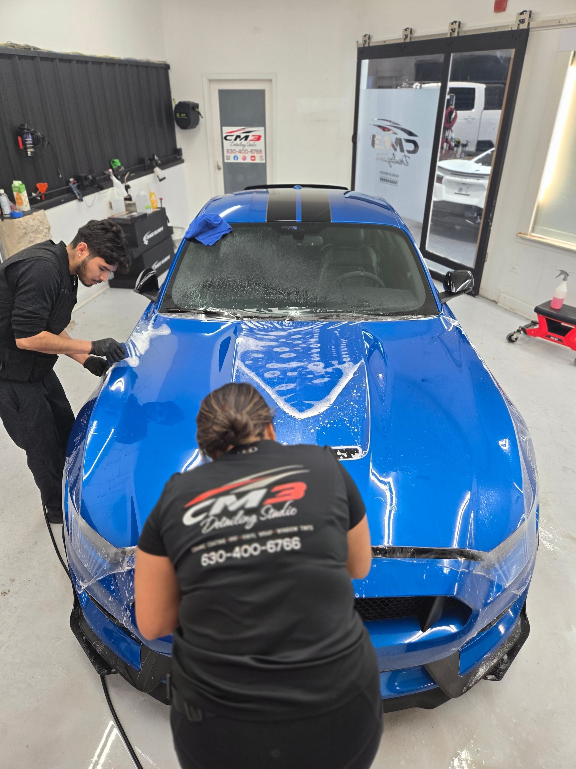 Two people applying protective film to a blue sports car in a shop.