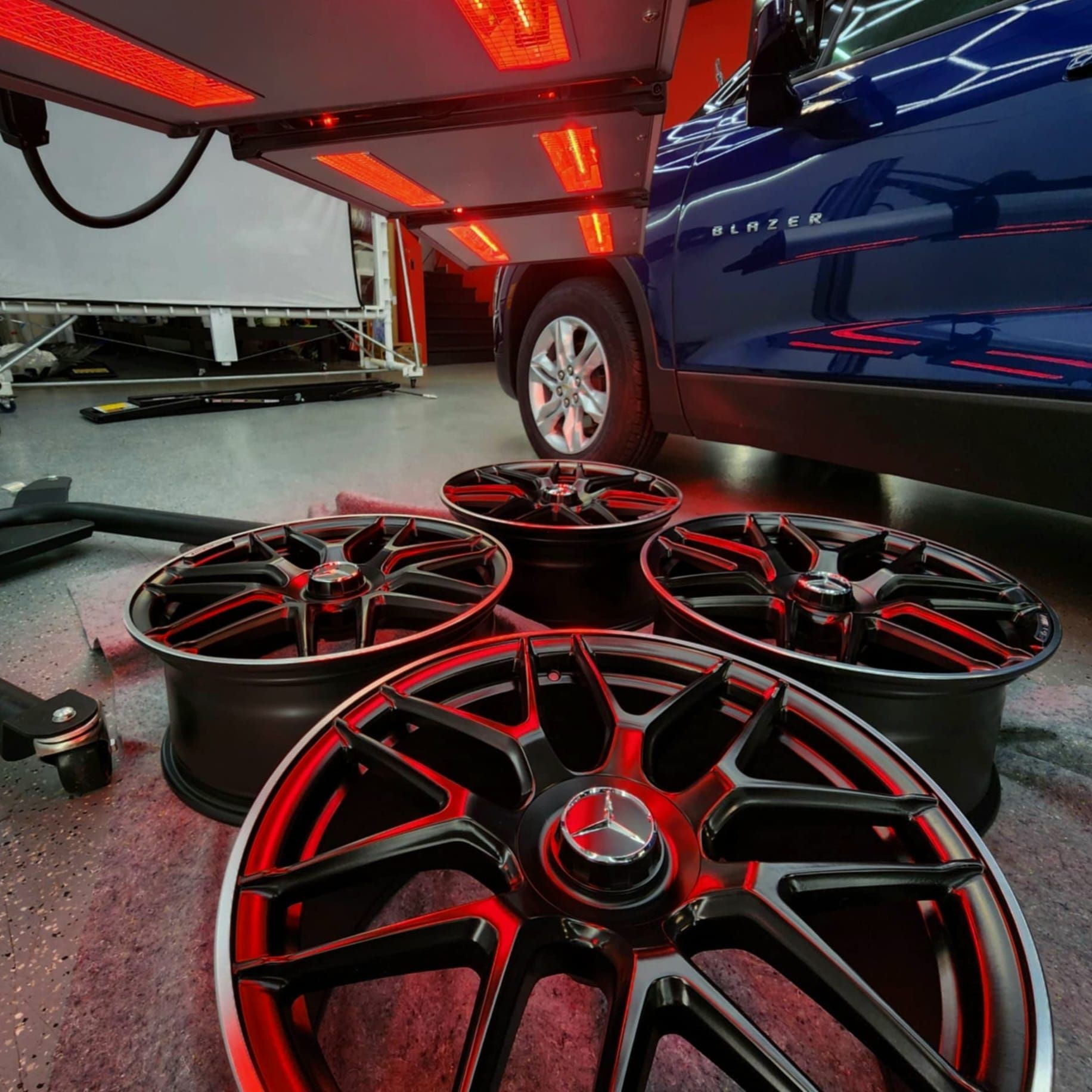 Four black and red car rims are on a garage floor near a blue car. Red heating lights are above.