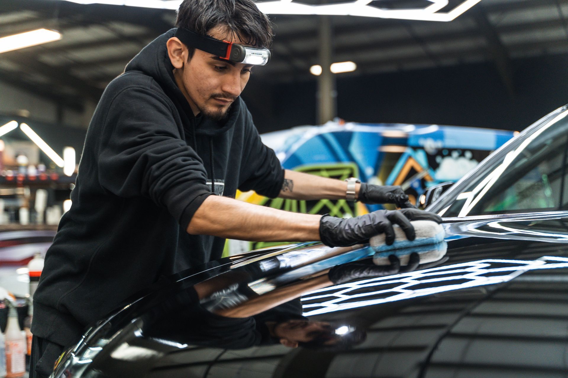 Man wearing headlamp polishes car's hood with a blue pad, in a garage.