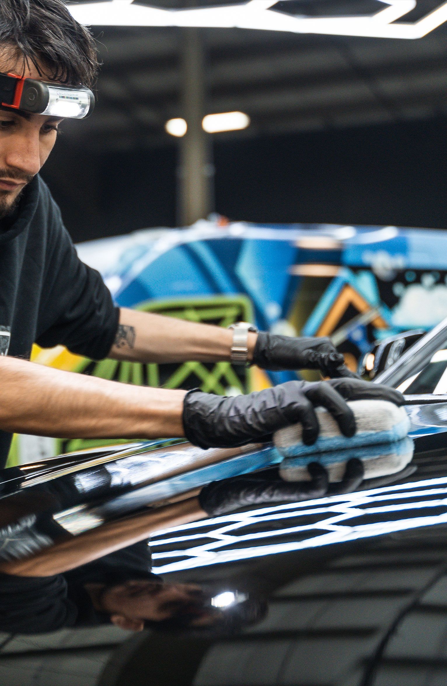 Man in black gloves polishes a car, using a buffing pad, under bright lights.