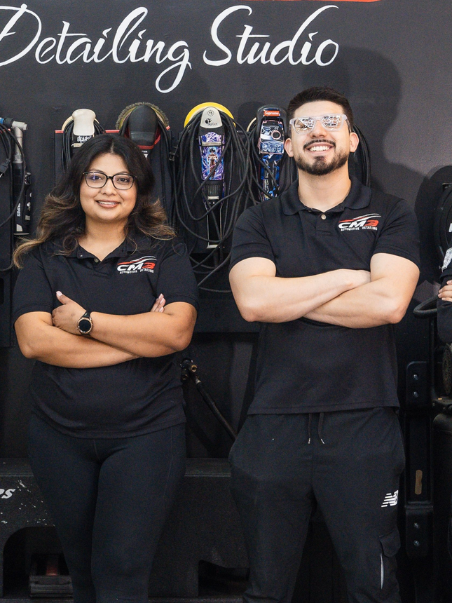 Two people wearing black logo shirts stand with arms crossed in front of a detailing studio wall with hanging tools.