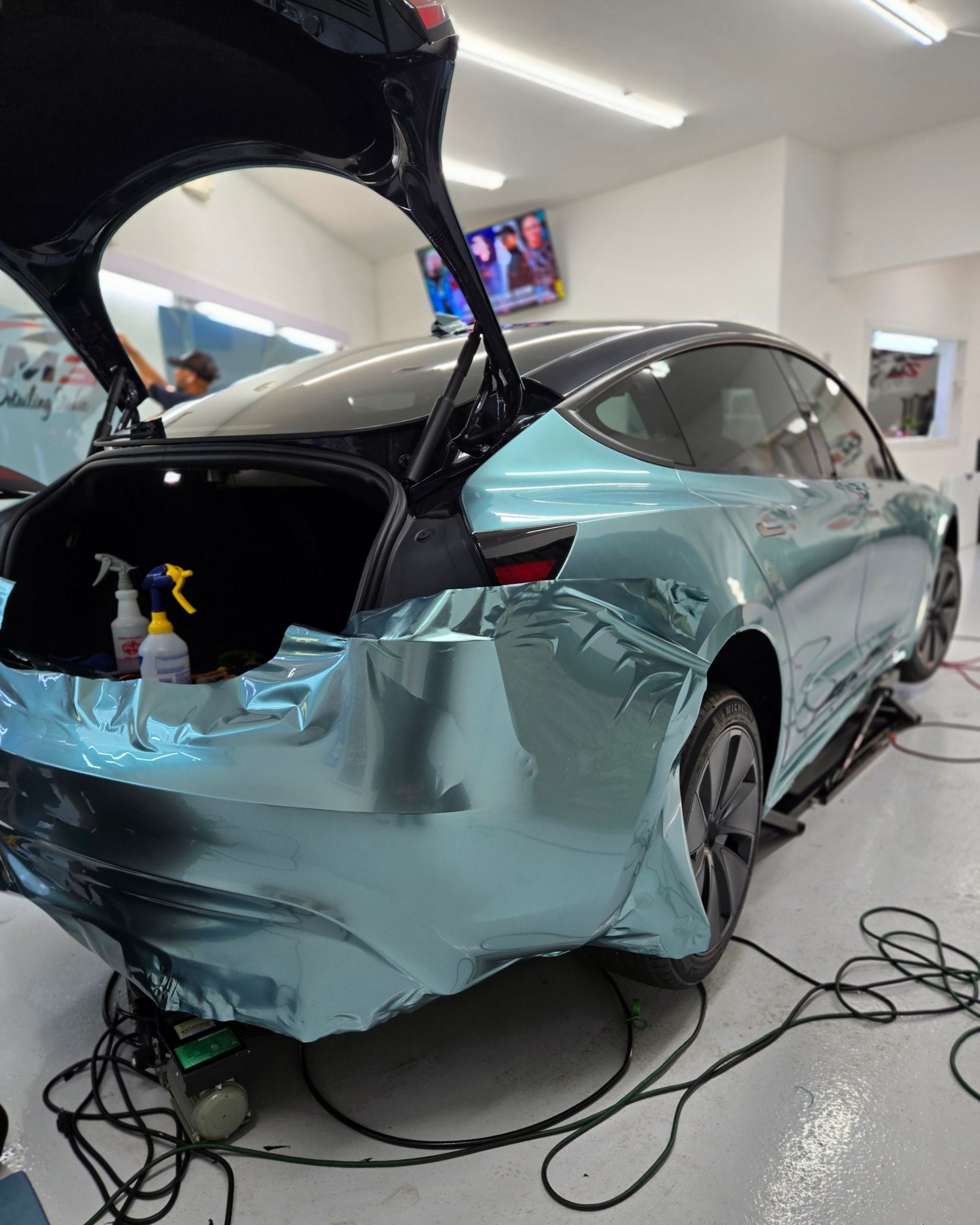 Person applying film to a car hood in a shop. The hood is gray.