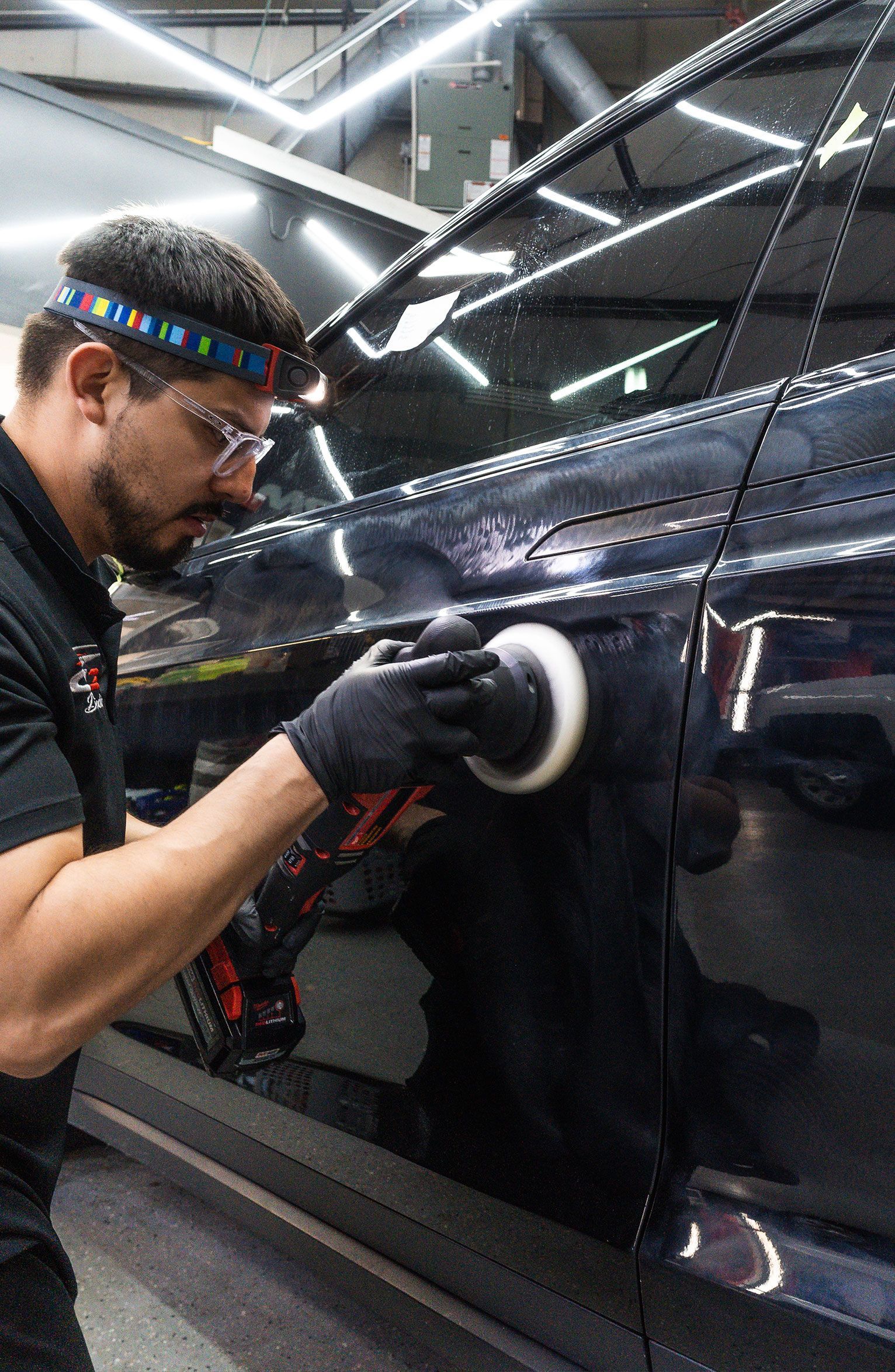 A person wearing safety glasses and a light polishes a black car door with a buffing pad.