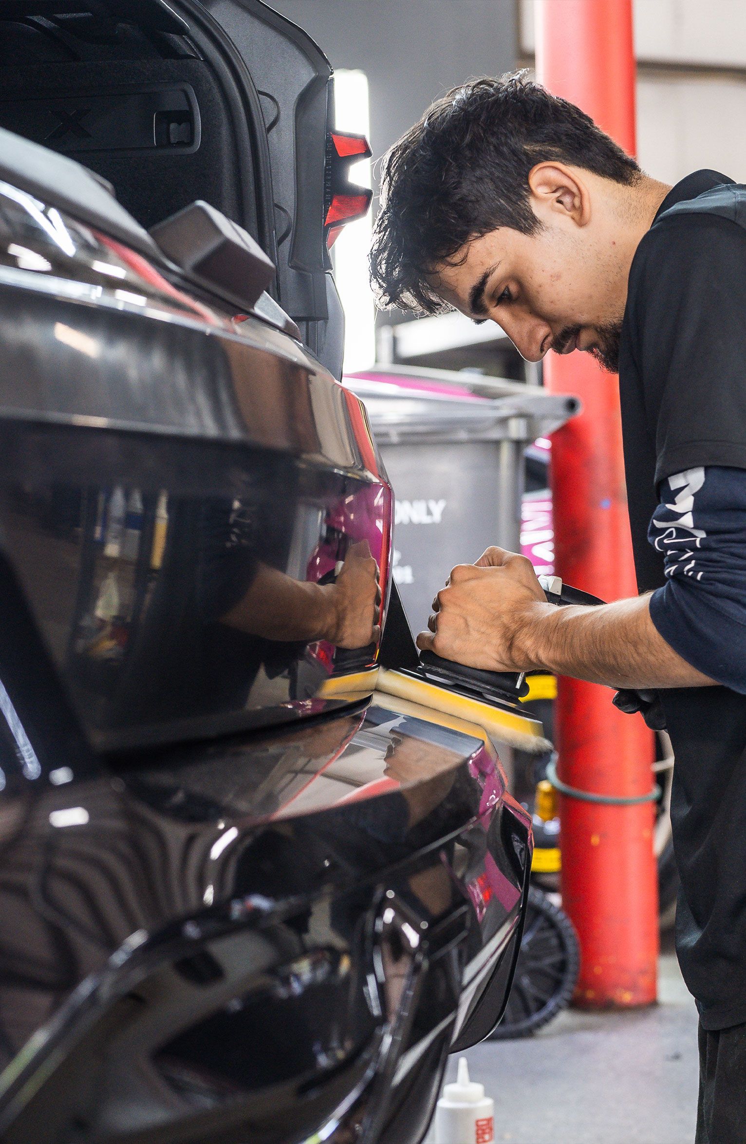 Man applying vinyl wrap to a black car's bumper in a garage using a tool.