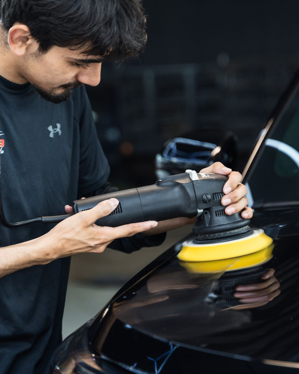 A professional in a black t-shirt uses a power polisher with a yellow pad to detail the hood of a black car.