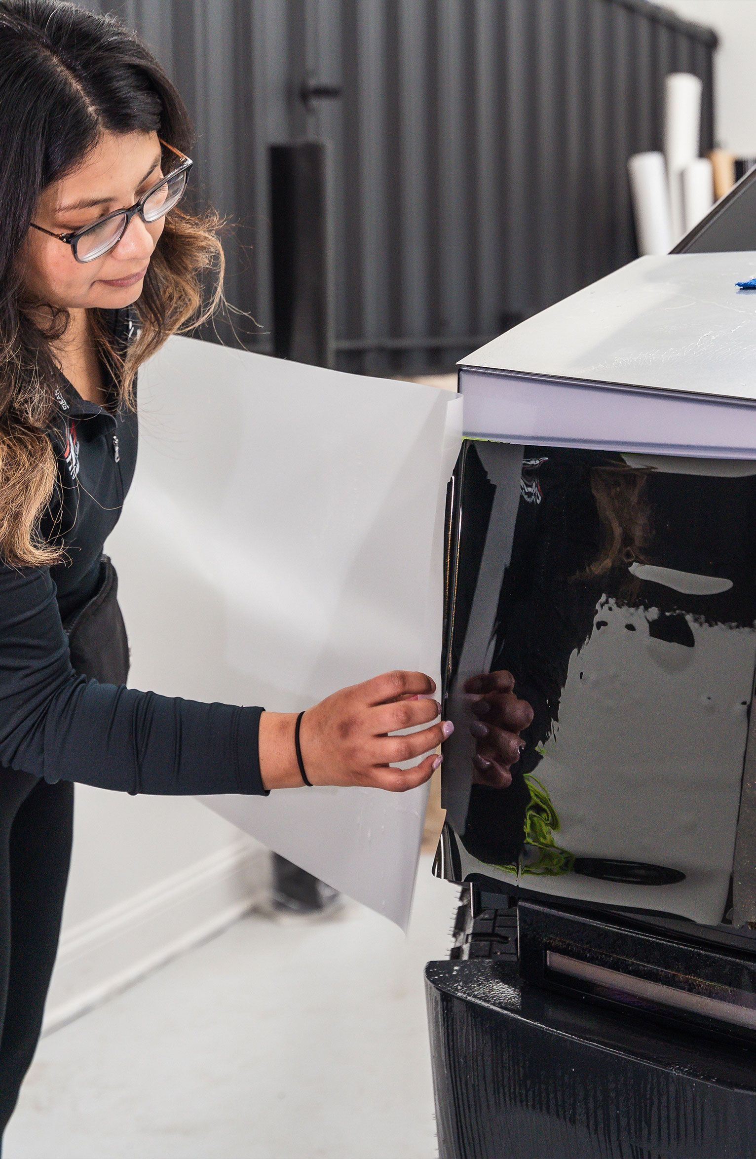 Woman applying white vinyl to a black car panel in a shop.