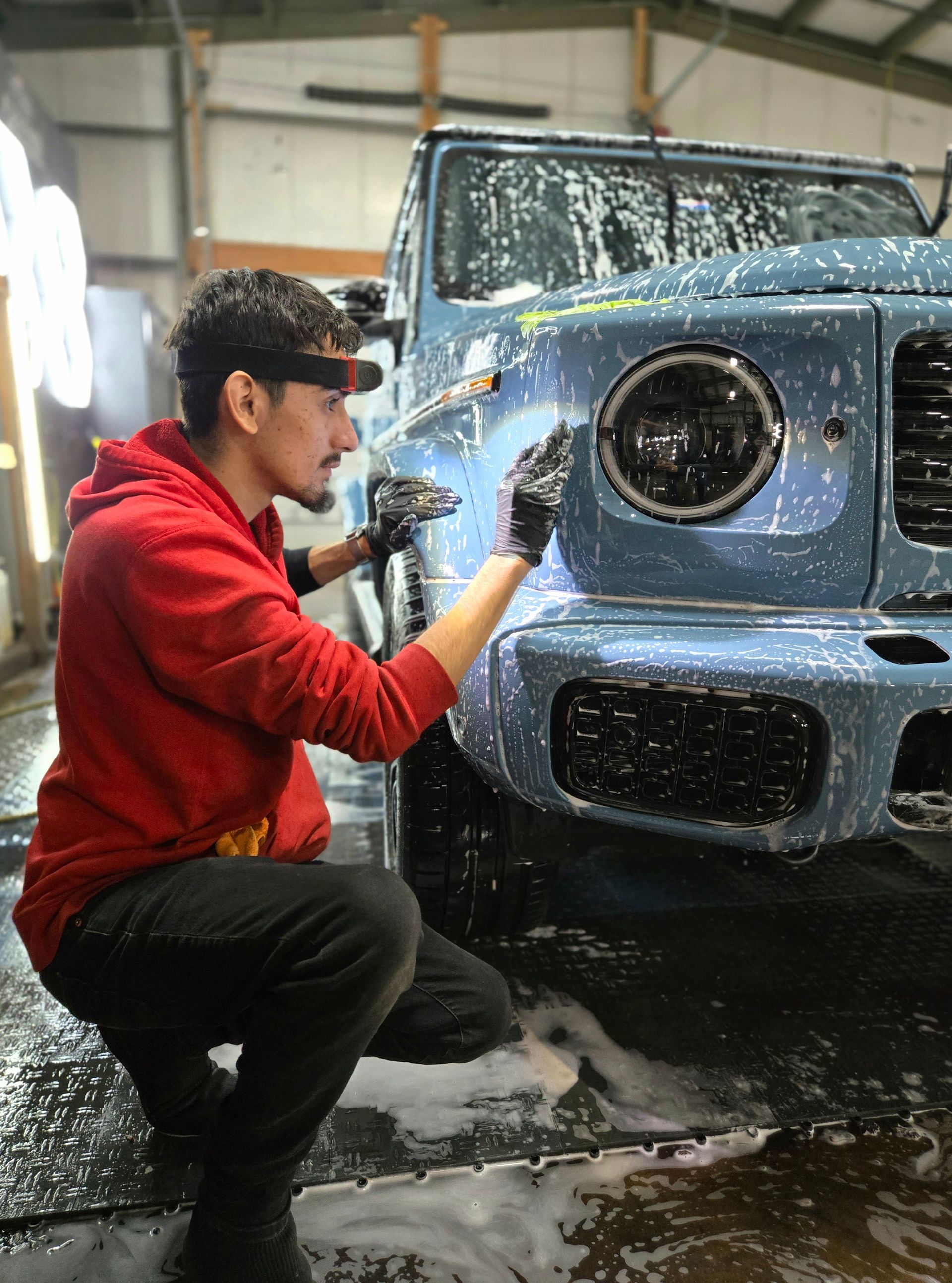 Man in red hoodie washing a light blue SUV with soapy water.