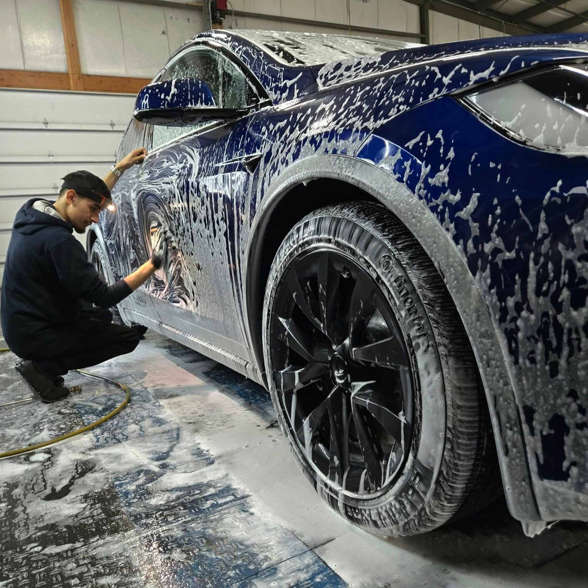 Person washes a blue car covered in soap, squatting near a black tire, in a garage.